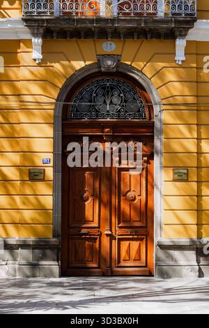 Vue frontale verticale d'une double porte en bois élaborée sertie dans une arche, sur une façade coloniale jaune vif avec des détails en fer forgé. Banque D'Images