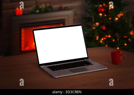 Ordinateur portable avec écran blanc sur table en bois, intérieur confortable avec arbre de Noël, tasse et cheminée incandescente en arrière-plan Banque D'Images