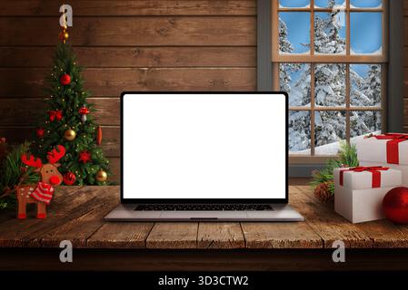 Ordinateur portable avec écran blanc vierge sur table en bois rustique, arbre de Noël, cadeaux et vue sur la forêt enneigée à travers la fenêtre de cabine en bois Banque D'Images