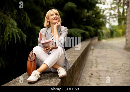 Une femme est assise confortablement sur un mur de pierre, lisant un livre et souriant. Elle profite d'une journée ensoleillée entourée d'arbres verts, créant un calme et réfléchissant Banque D'Images
