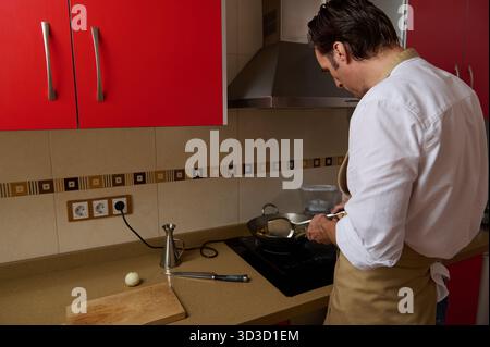 Un homme dans un tablier cuisine à un poêle dans une cuisine rouge vif, remuant une casserole pendant qu'il prépare le dîner avec l'aide d'un membre de la famille à proximité. Banque D'Images