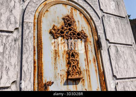 Une croix de fer forgé très rouillée et ornée sur la porte d'un mausolée vieilli, capturant texture et pourriture au cimetière de Prazeres. Banque D'Images