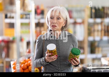 Shopping dans le supermarché de produits - acheteur fremale mature fait un choix lors de l'achat d'olives en conserve Banque D'Images
