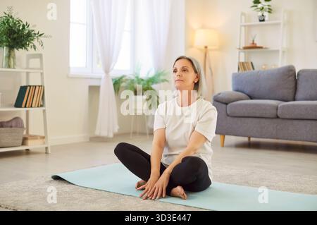 Femme senior faisant du yoga à la maison et assise dans la pose à jambes croisées sur le tapis d'exercice Banque D'Images