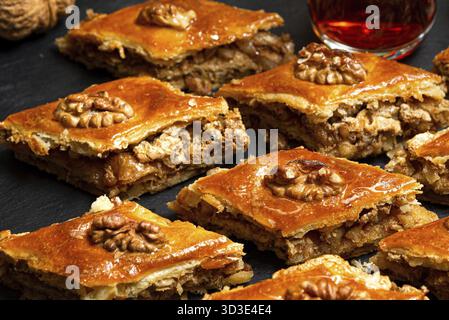 Nourriture, baklava au miel avec des noix sur ardoise noire. Portions de bonbons orientaux. Morceaux sucrés turcs. Thé versé dans le verre d'armudu en arrière-plan Banque D'Images