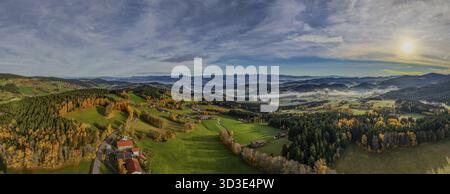 L'image montre un vaste paysage verdoyant avec des collines et des forêts. Le soleil se lève et illumine les feuilles d'automne colorées. Le brouillard balaye le val Banque D'Images