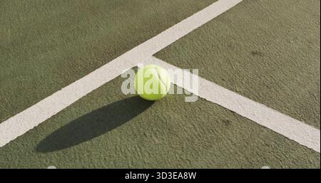 Balle de tennis reposant projetant une ombre allongée sur un terrain vert dur, avec des lignes de limite blanches nettes Banque D'Images