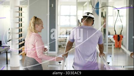 Homme marchant en tee-shirt violet utilisant des barres parallèles dans la salle de rééducation, thérapeute guidant avec harnais orange Banque D'Images