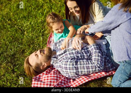 Famille heureuse allongée sur une couverture à l'extérieur, riant, les enfants chatouillant père, souriant Banque D'Images