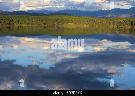 Un lac clair reflétant parfaitement les montagnes enneigées et une dense forêt de pins verts sous un ciel bleu nuageux. Un paysage vibrant et superposé idéal pour Banque D'Images