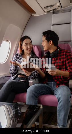 Un jeune couple asiatique assis dans un avion, souriant tout en tenant des passeports et un smartphone. Ils semblent excités et prêts pour leur voyage, dresse Banque D'Images