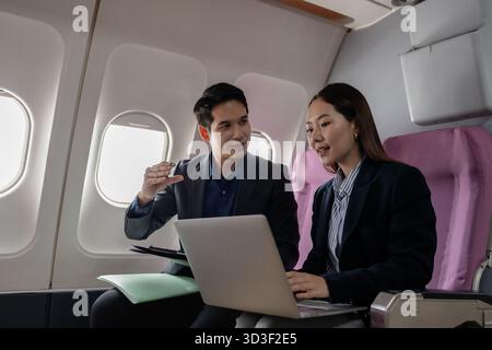 Professionnels asiatiques travaillant ensemble sur un ordinateur portable tout en étant assis sur un avion commercial. Ils semblent discuter d'un projet ou d'un présentati Banque D'Images
