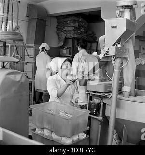 Une photo d'archives des années 1970 montre l'arrière-boutique d'une épicerie dans le microdistrict de Cherevkovka, Sloviansk, RSS d'Ukraine. Une employée, vêtue d'un manteau blanc et d'un foulard, exploite une machine d'emballage. Elle utilise une pelle pour répartir les marchandises en vrac (peut-être du gruau ou du sucre) d'une trémie dans des sacs en papier, qui sont dans un bac en plastique ci-dessous. En arrière-plan, d'autres membres du personnel travaillent, et de grands sacs sont empilés haut. Cette scène capture le processus de pré-emballage dans un magasin soviétique typique. C’est un moment de travail quotidien dans le Donbass pacifique avant la guerre. Banque D'Images