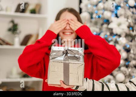 Une belle jeune femme reçoit une boîte cadeau sur fond lumineux d'arbre de Noël. La femme couvrit ses yeux avec ses mains, la main devant son ho Banque D'Images