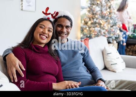 Couple diversifié assis sur le canapé, portant serre-tête en bois et chapeau de Père Noël à côté de l'arbre de Noël dans la chambre Banque D'Images