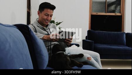 Tablette navigation homme asiatique portant un sweat à capuche se prélassant sur un canapé en velours marine dans le salon, espace de copie Banque D'Images