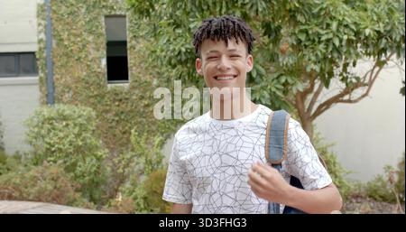 Garçon adolescent souriant debout dans la cour du campus portant un sac à dos en denim et portant un T-shirt à motifs Banque D'Images