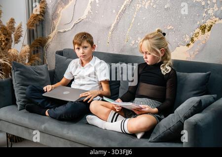 Canapé pour ordinateur portable enfants - deux enfants assis sur un canapé tandis que l'un utilise un ordinateur portable et l'autre lit. Banque D'Images