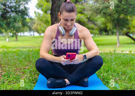 Femme sportive assise sur le tapis de yoga à l'aide d'un smartphone à l'extérieur dans un parc vert Banque D'Images