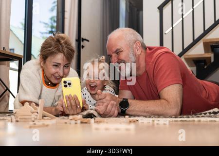 Des grands-parents affectueux et leur petite-fille joyeuse sourient alors qu'ils explorent un smartphone ensemble. La famille se lie sur des moments partagés dans une maison confortable Banque D'Images