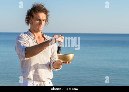 Un homme pratique la méditation en utilisant des bols chantants tibétains au bord de la mer, en se concentrant sur l'harmonie et la pleine conscience. L'océan serein en toile de fond renforce l'apaisement Banque D'Images
