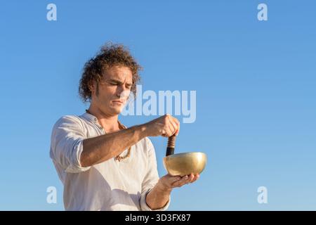Un homme médite en utilisant un bol chantant tibétain dans un environnement extérieur serein, en se concentrant sur la pleine conscience et la détente. Cette pratique améliore le spirituel Banque D'Images
