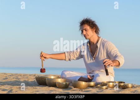 Un homme médite au bord de la mer en utilisant des bols chantants tibétains, favorisant la relaxation et la pleine conscience. La pratique est renforcée par la toile de fond apaisante de l'océan, Banque D'Images
