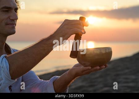 Un homme médite au coucher du soleil en utilisant un bol chantant tibétain au bord de la mer. Cette pratique, connue pour ses vibrations apaisantes et ses bienfaits de thérapie sonore, s’améliore Banque D'Images