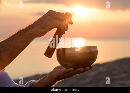 Une scène sereine d'un homme utilisant un bol chantant tibétain pendant la méditation au bord de la mer. Le bol est élevé à la main, capturant le coucher de soleil dans le dos Banque D'Images