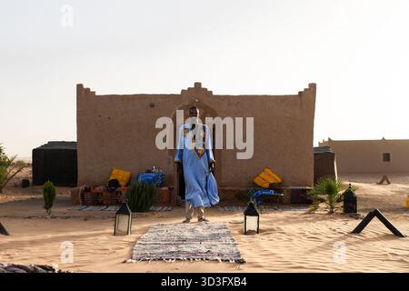 Un homme nomade vêtu de vêtements traditionnels se tient devant un camp du désert du Sahara à Merzouga, au Maroc. Le soleil couchant projette des tons chauds, mettant en évidence Banque D'Images