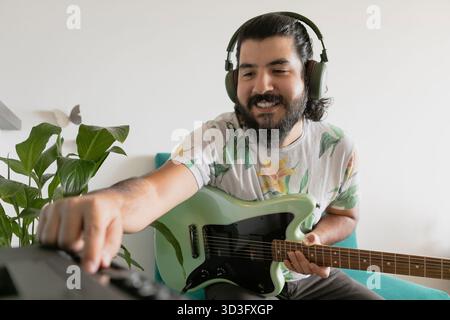 Homme souriant portant des écouteurs tout en jouant d'une guitare électrique et en réglant les commandes sur un mélangeur de musique à la maison, appréciant la créativité, la conception sonore, un Banque D'Images