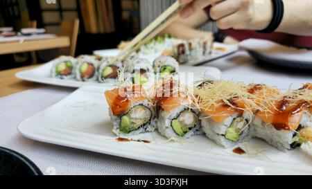Une image en gros plan présentant une délicieuse tartinade à sushis, avec des rouleaux recouverts de saumon frais et d'avocat. Une main utilise habilement des baguettes à sele Banque D'Images