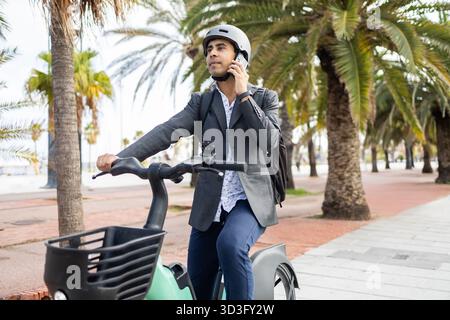 Homme hispanique conduit un e-bike sur un chemin bordé de palmiers, portant un casque et parlant sur son smartphone. Engagé dans un appel, il équilibre les déplacements urbains et Banque D'Images