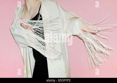 Femme recadrée méconnaissable portant une veste élégante avec des pompons, dansant gracieusement devant un fond rose, mettant en valeur le mouvement et la mode. Banque D'Images