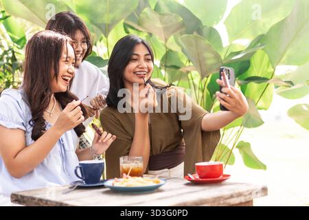 Un groupe d'adolescents asiatiques joyeux s'amuse dans un café indonésien, prenant des selfies et sirotant des boissons. L'atmosphère vibrante améliore leur Banque D'Images