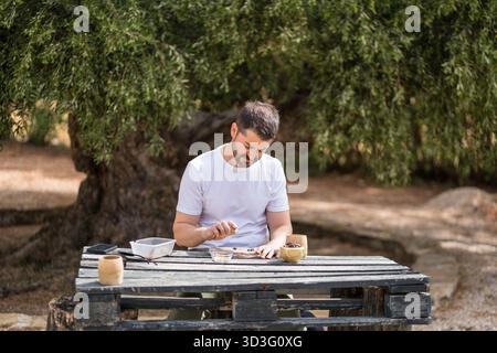 Un homme en chemise blanche est assis à une table rustique en plein air, craquant méticuleusement les noix de pin. Entouré par la nature, il se concentre sur l'extraction du nutritif Banque D'Images