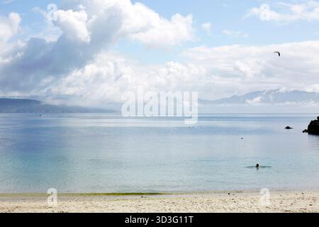 Une scène de plage paisible en Galice, Espagne, avec des eaux calmes et des montagnes lointaines sous un ciel nuageux. Une personne profite d'une baignade tranquille, entourée par Banque D'Images