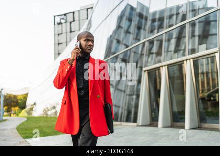 Un jeune homme LGBTQ marche en toute confiance devant un bâtiment moderne en verre. Il porte une veste rouge, parle sur son téléphone, et porte un ordinateur portable, symb Banque D'Images