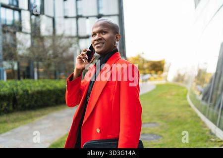 Un jeune homme LGBTQ dans une veste rouge éclatante se tient en toute confiance à l'extérieur, parlant au téléphone avec le sourire. L'arrière-plan présente une architecture moderne Banque D'Images