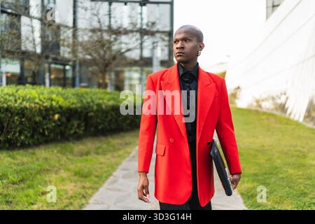 Un jeune homme LGBTQ stylé sort en toute confiance, portant un blazer rouge frappant, placé sur un fond urbain moderne avec des espaces verts. Banque D'Images