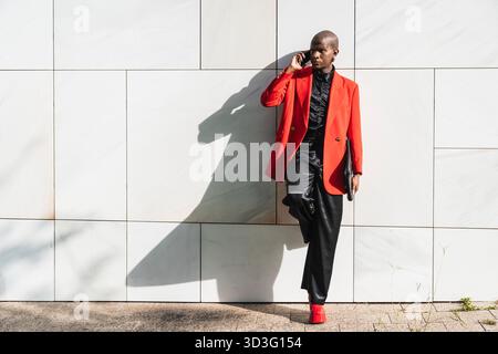 Un jeune homme LGBTQ portant une veste rouge vif s'appuie contre un mur carrelé moderne tout en parlant sur son téléphone portable. Il respire la confiance et le style, showca Banque D'Images
