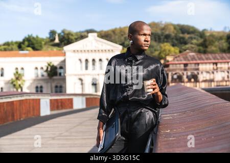 Un jeune homme LGBTQ dans une tenue élégante profite d'un café à l'extérieur, tenant une tasse avec un regard contemplatif. L'arrière-plan présente une architecture classique BA Banque D'Images