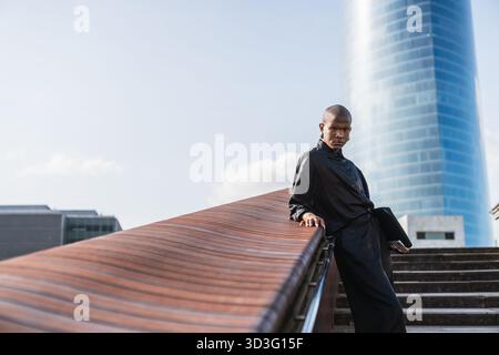Un jeune homme LGBTQ se tient confiant dans un environnement urbain moderne. Il s'est positionné sur une structure architecturale élégante, dégageant style et calme Banque D'Images