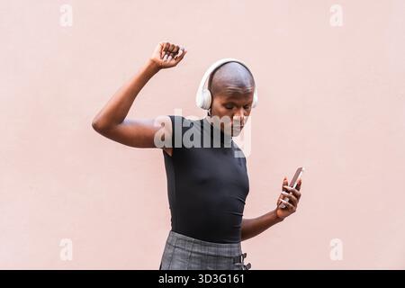 Un jeune homme LGBTQ aime la musique sur ses écouteurs, perdu dans l'instant présent. Il se tient confiant sur un fond rose minimaliste, incarnant la liberté et Banque D'Images