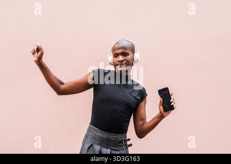 Jeune homme LGBTQ danse joyeusement au son de la musique avec des écouteurs, tenant un smartphone. Son expression respire le bonheur et la liberté contre un dos neutre Banque D'Images