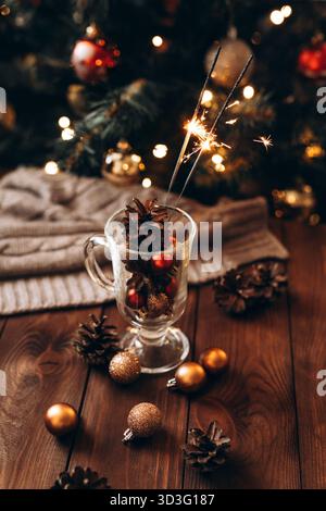 Scène de Noël confortable avec des paillettes et des pommes de pin dans une tasse en verre, serrée contre un sapin magnifiquement éclairé avec des ornements dorés, capturant la fête Banque D'Images