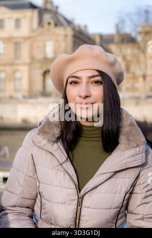 Une femme est capturée à Paris portant un béret beige et une veste d'hiver. Sa tenue élégante complète l'architecture parisienne classique dans le backgr Banque D'Images