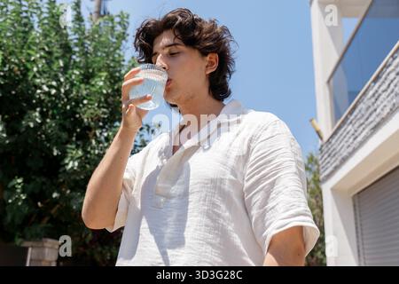 Un jeune homme en chemise blanche boit de l'eau dans un verre sous un ciel bleu clair. La perspective à angle bas souligne la chaude journée d'été et son rafraîchissement Banque D'Images