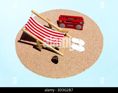 Vue de dessus d'une scène de plage miniature animée avec une chaise longue rayée rouge et blanche, radio rouge, pantoufles blanches et une boisson rafraîchissante sur un sable Banque D'Images