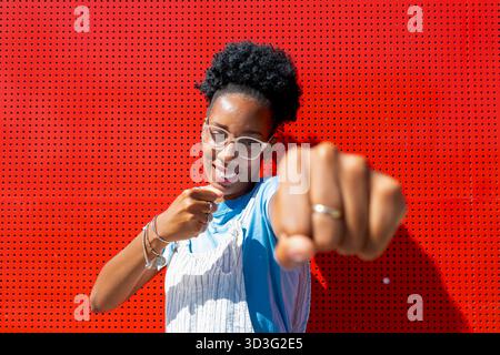 Une femme souriante aux cheveux bouclés et aux lunettes pose énergiquement devant un mur rouge vif. Son poing est levé vers la caméra, exsudant la confiance Banque D'Images
